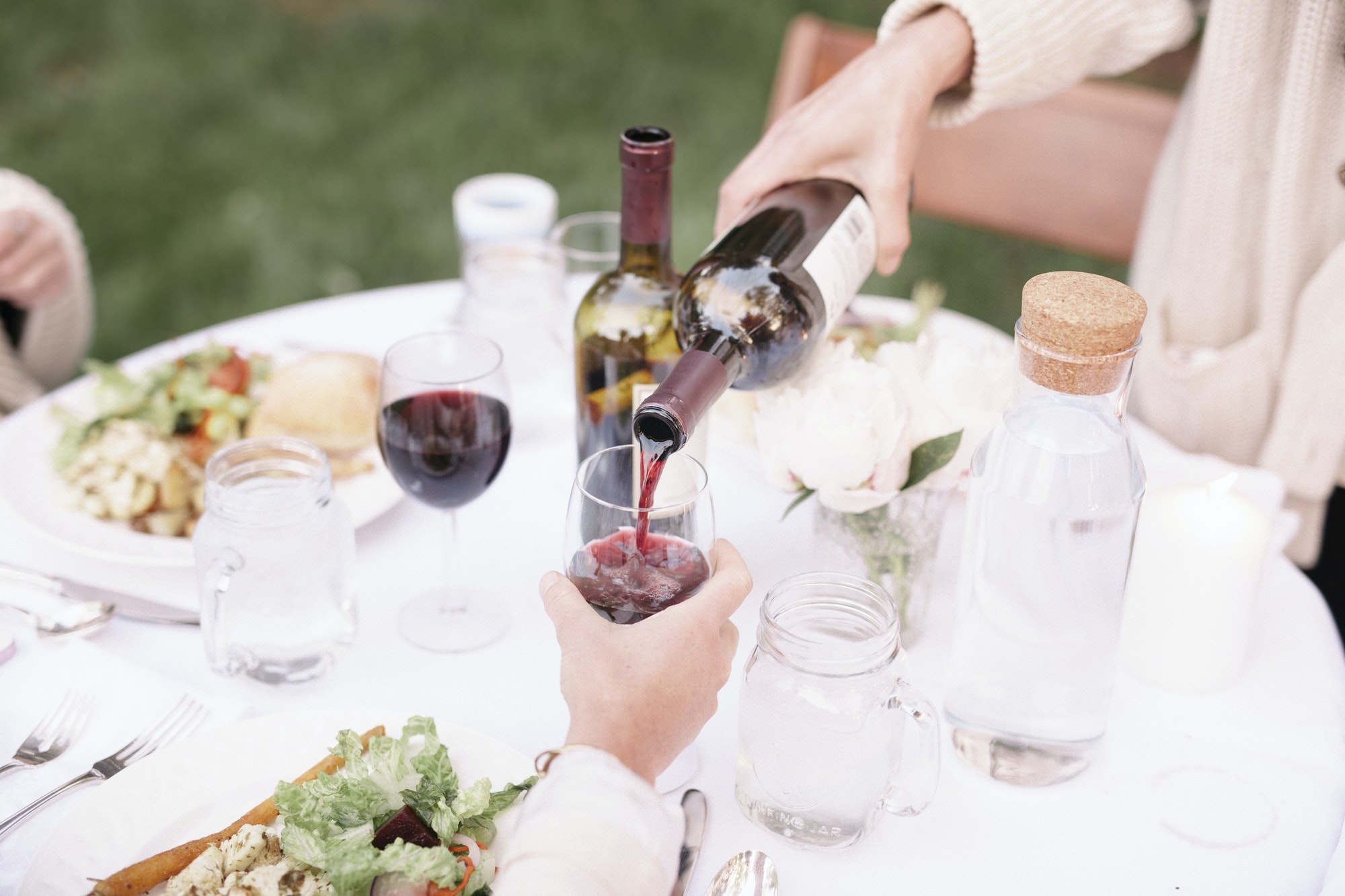 Close up of a a table with food and drink in a garden, woman pouring a glass of red wine.
