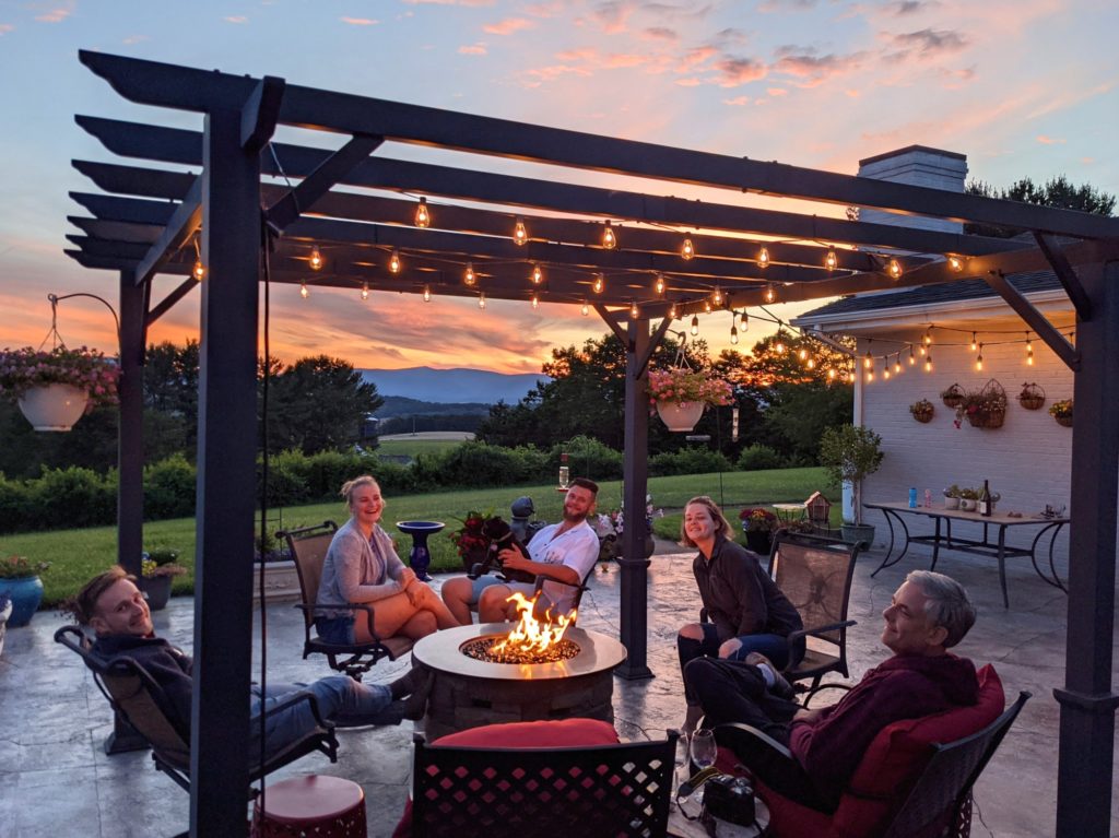 A family relaxing around a fire table in outdoor seating area under pergola with lights at sunset